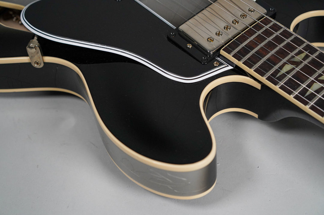 Close-up of a black guitar body with cream binding, showing a pickup, part of the fretboard with parallelogram inlays, and light aging.