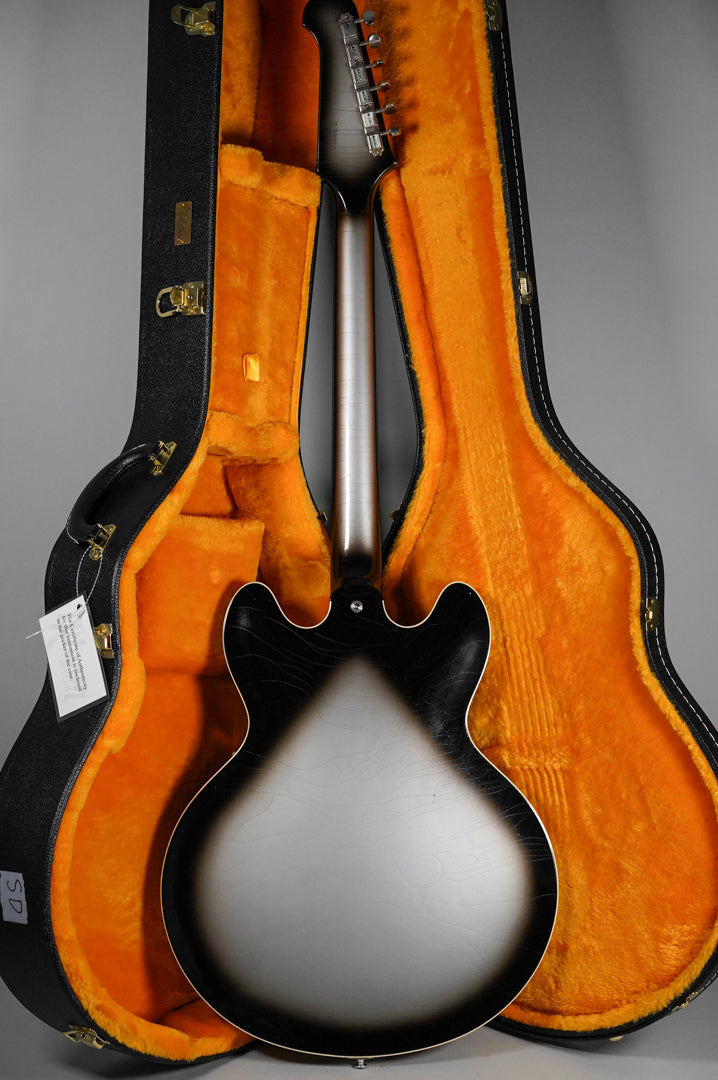 The back of a silverburst semi-hollow guitar with light aging, showing the neck and headstock, resting in an open orange-lined case.