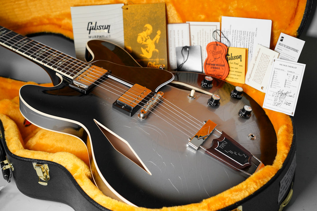 A Silverburst semi-hollowbody electric guitar with f-holes, light aging, and a Trini Lopez tailpiece, resting in a yellow-lined hard case.