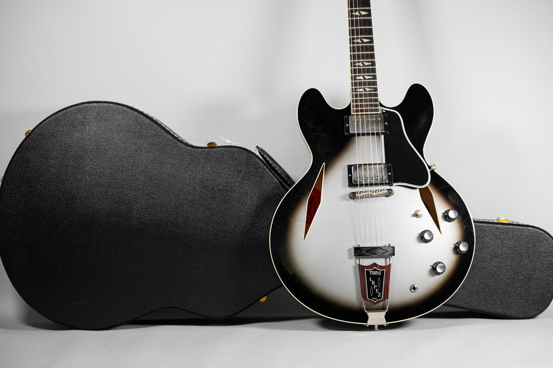 Silverburst semi-hollow guitar with black pickguard, two humbuckers, and four control knobs, leaning against its open black hard case.