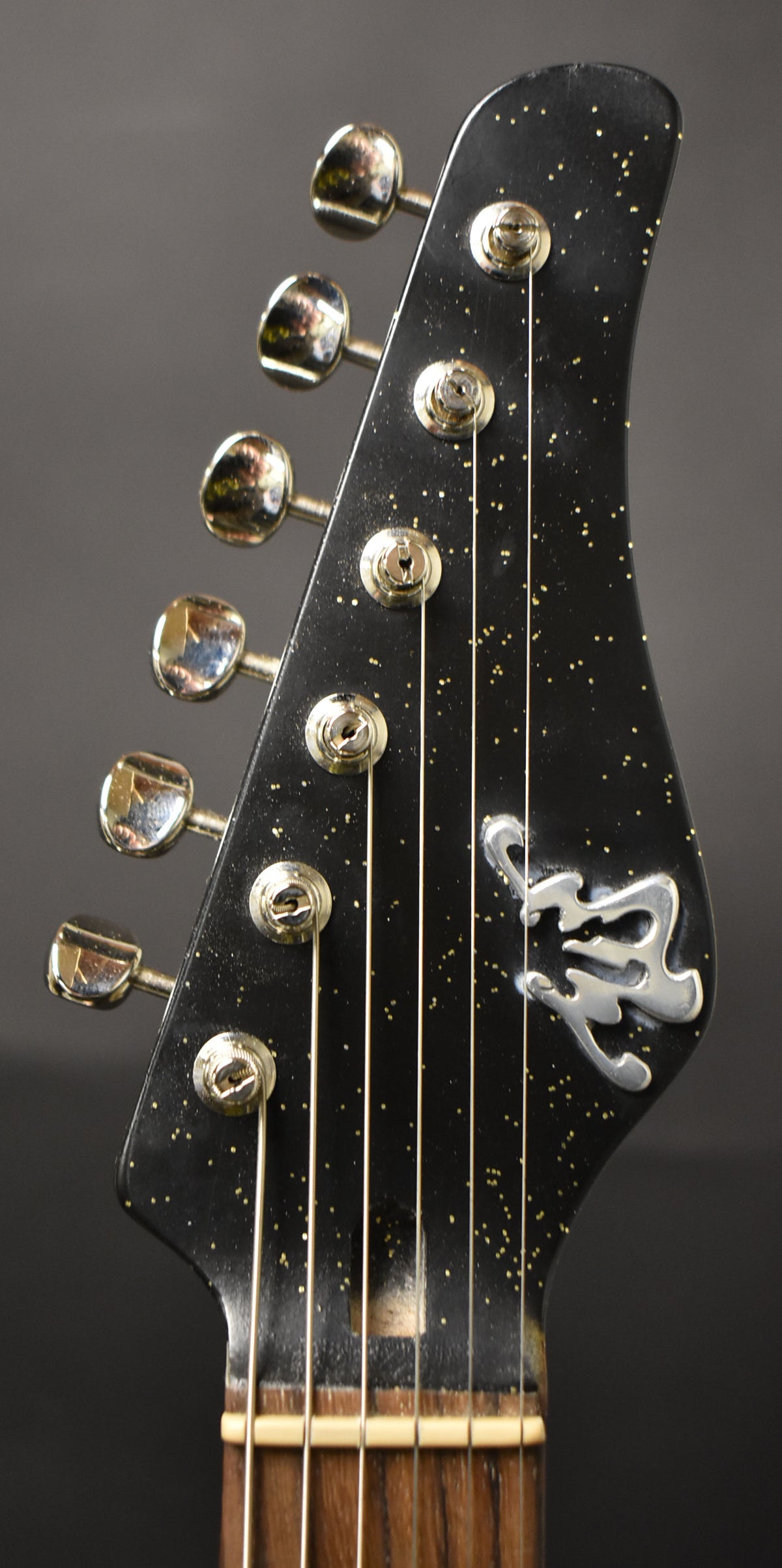 Close-up of the black sparkle headstock with six chrome tuners, a silver logo, and a visible nut and fretboard.