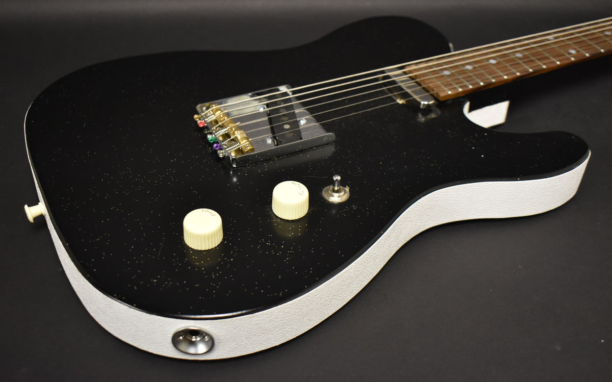 The black sparkle electric guitar, with white textured sides, is angled to show its two cream knobs and chrome bridge with colorful string ends.