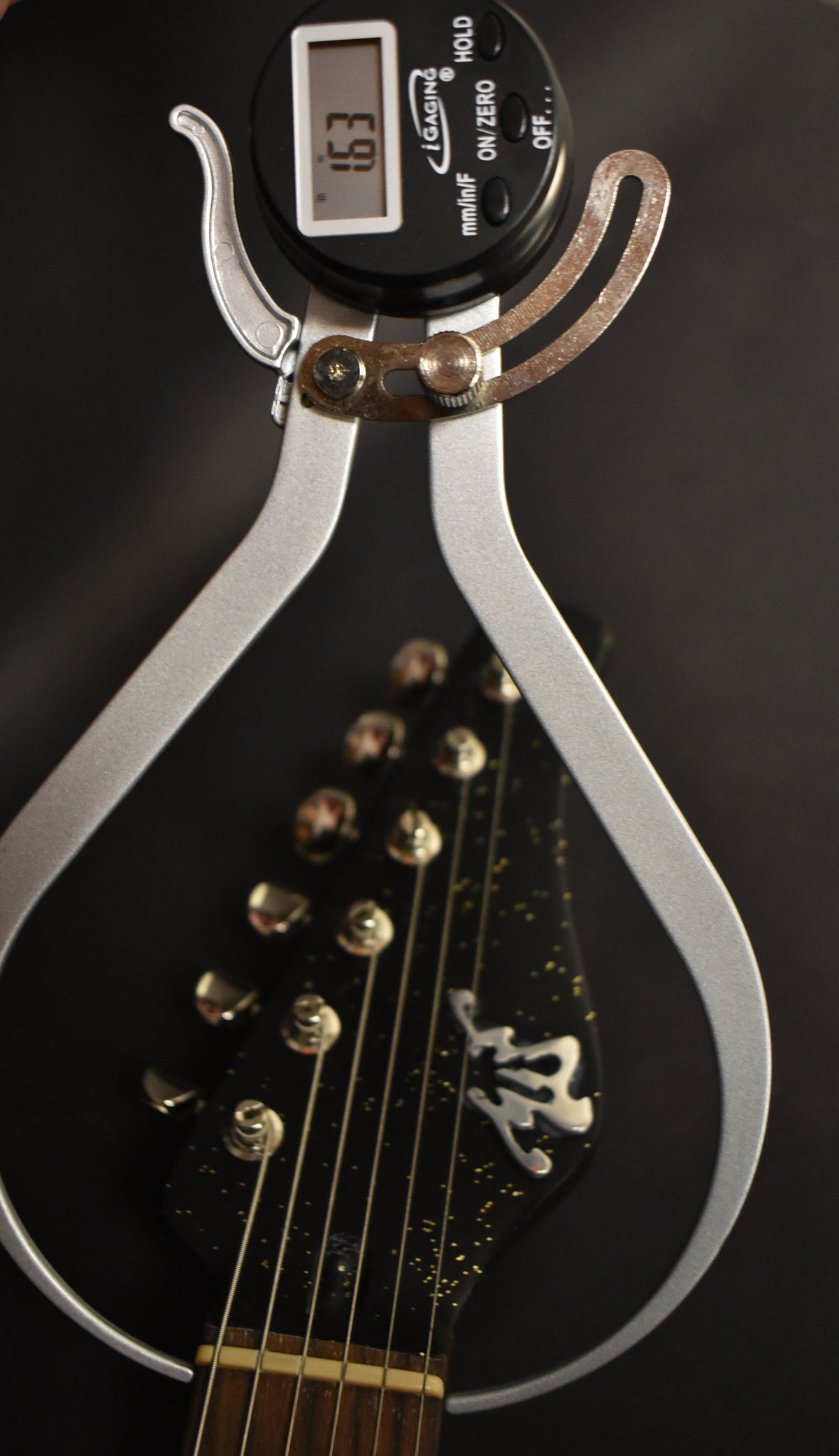 A digital caliper measuring 1.63 inches is clamped over the black sparkle headstock with gold flecks, showing six tuners.