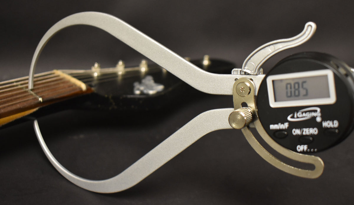 A digital caliper measures the guitar's nut width, showing "0.85" on its screen, with the fretboard visible behind it.