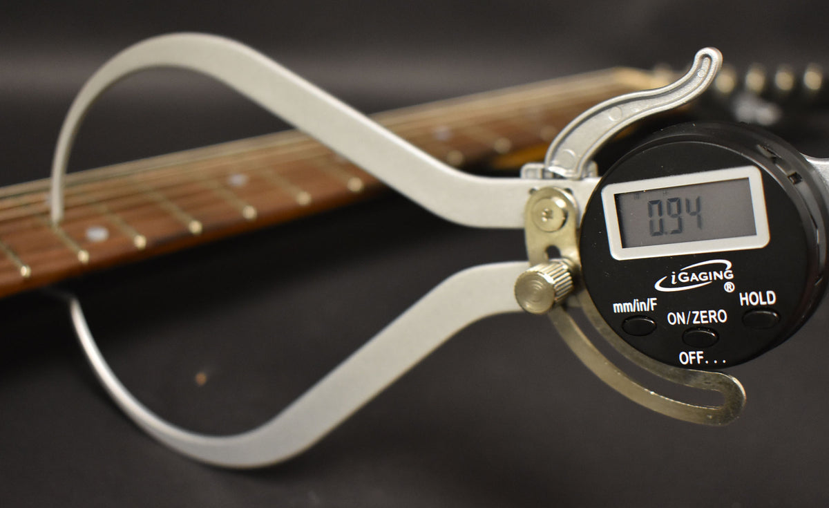A digital caliper measures the guitar's fretboard, showing a reading of 0.94, with the neck angled away from the viewer.