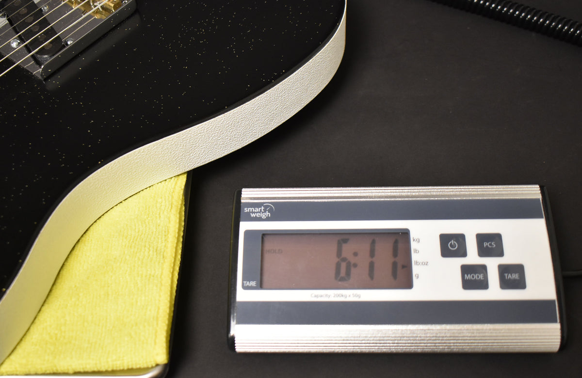 The guitar's black sparkle body with cream binding rests on a yellow cloth, next to a digital scale displaying "6:11".