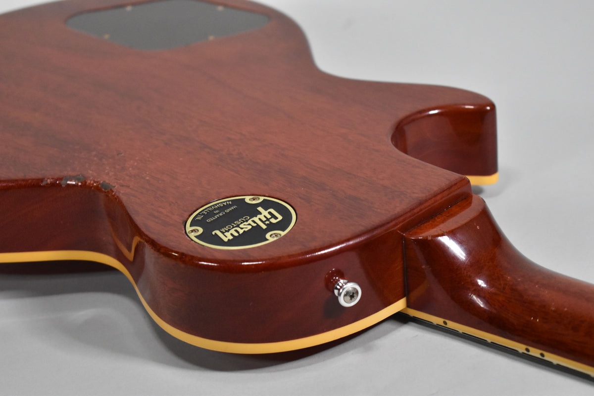 The back of the cherry-finished guitar body, showing the neck joint, output jack, and a black circular backplate with gold lettering.