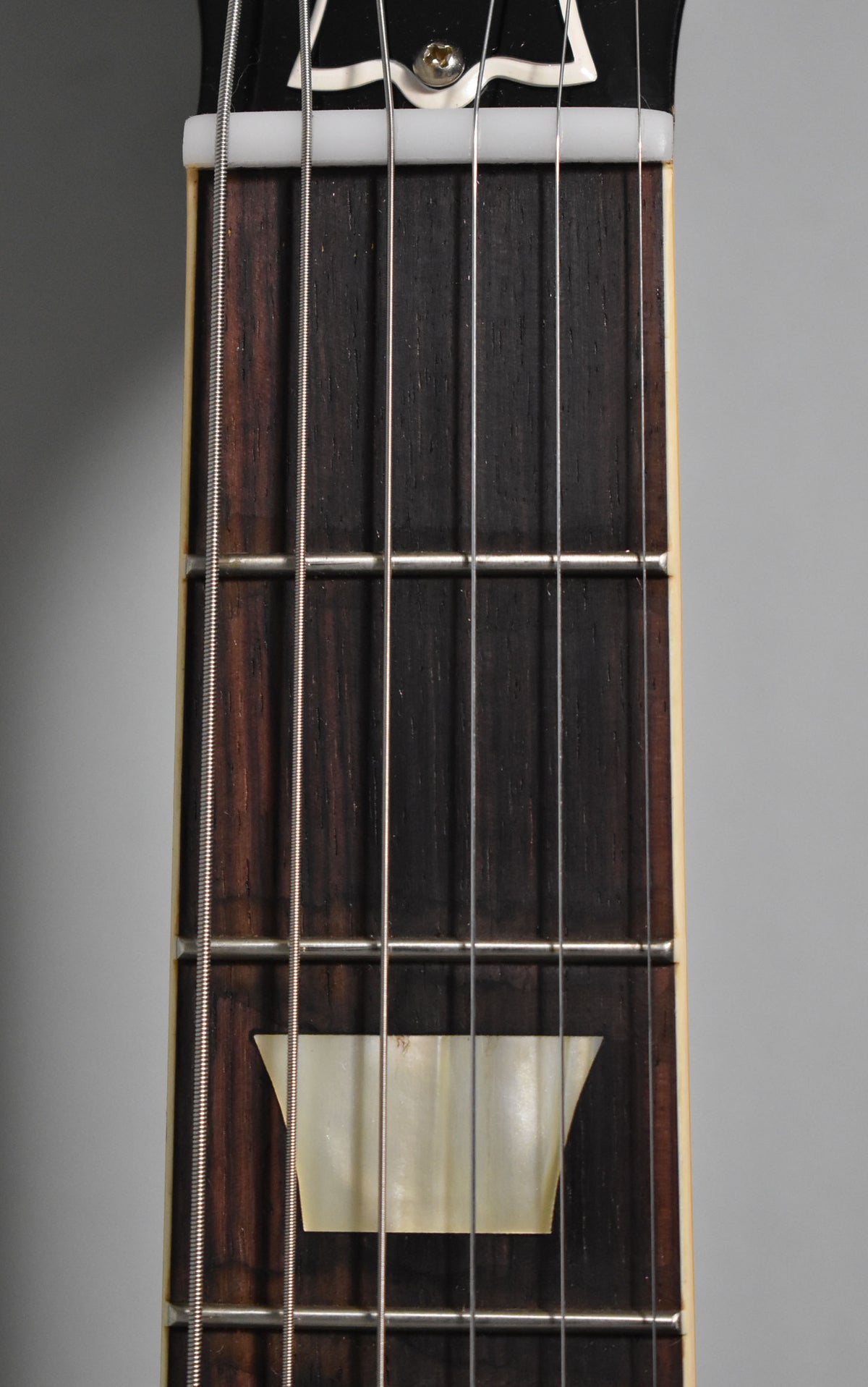 Close-up of the dark rosewood fretboard, showing the white nut, metal frets, and a trapezoid mother-of-pearl inlay.