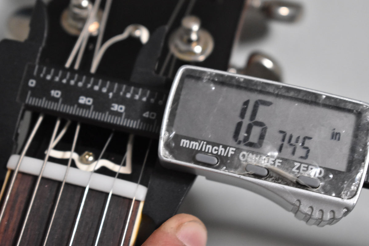 A close-up of the guitar's black headstock and nut, with a digital caliper measuring the nut width at 1.6745 inches.