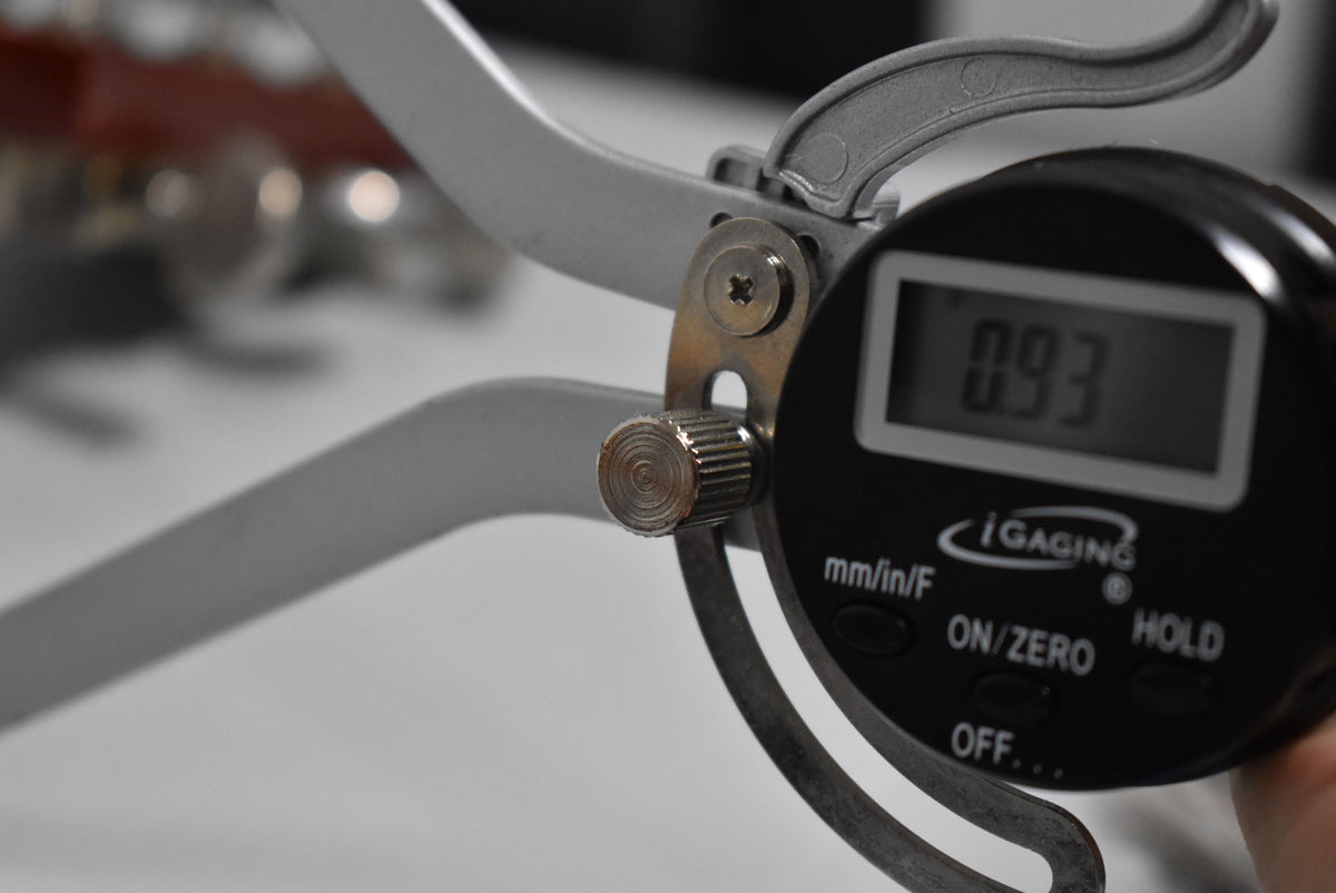 A digital caliper measures the guitar's nut width, displaying "0.93" on its screen, with the guitar blurred in the background.