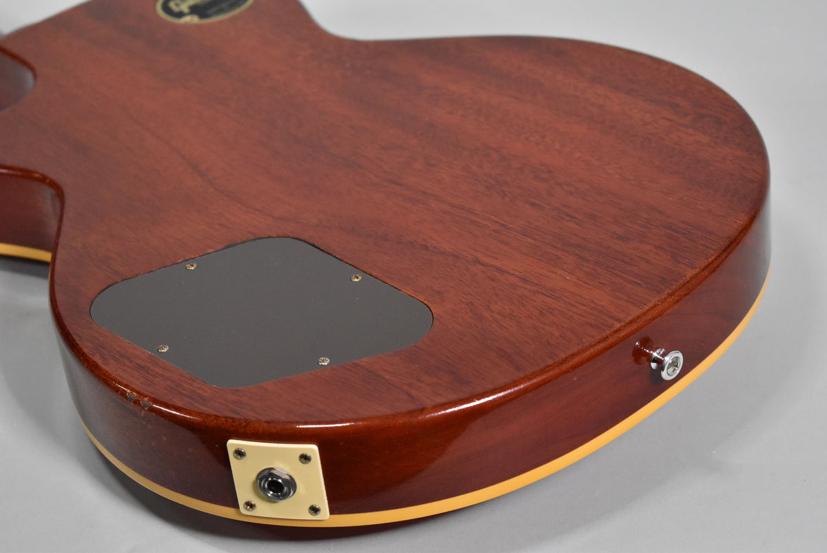 The back of a cherry-stained guitar, angled from the lower bout, showing the output jack, control cavity cover, and a strap button.
