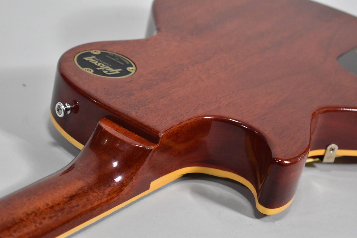 The back of a cherry-finished electric guitar, showing the neck joint, strap button, and a black oval Gibson Custom Shop decal.