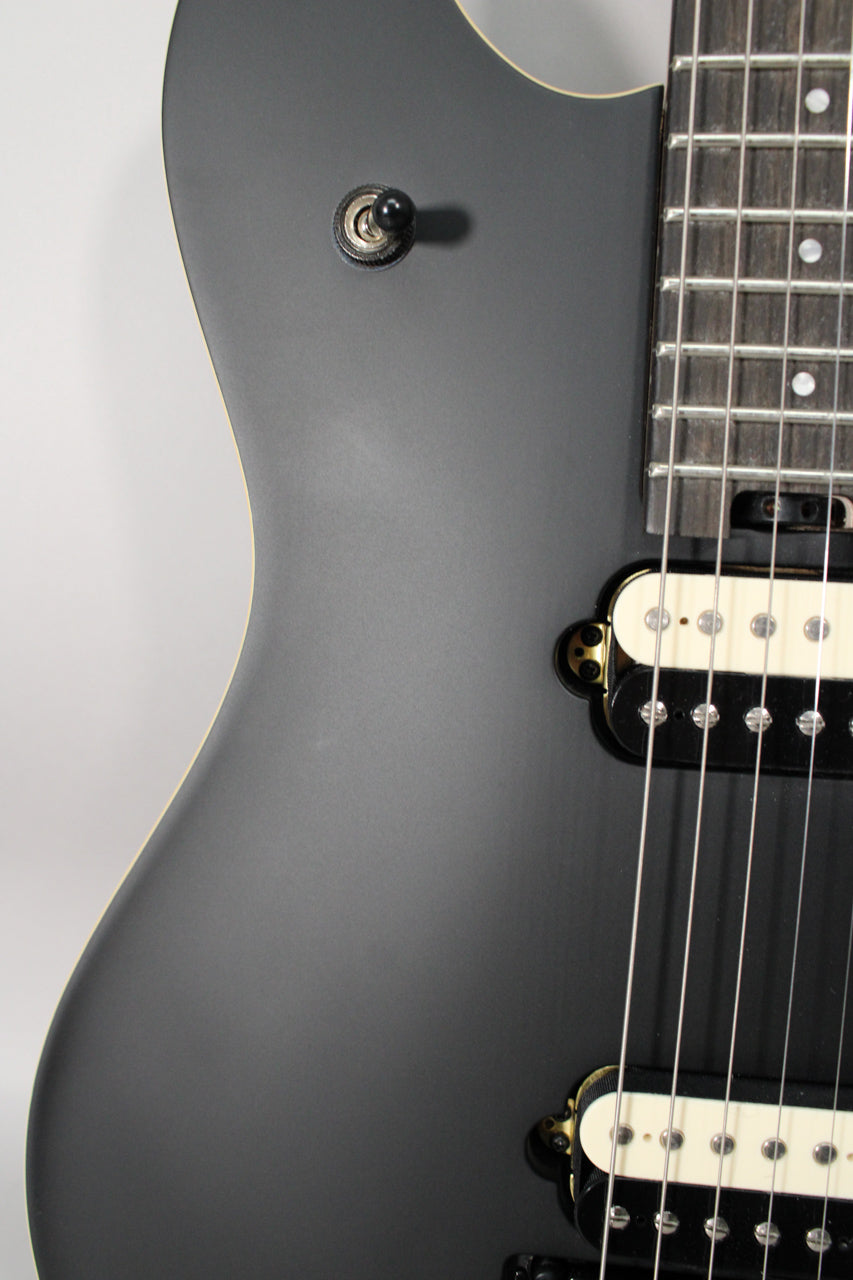 Close-up of the matte black body, showing the upper horn, a black toggle switch, and two humbucker pickups with cream and black bobbins.