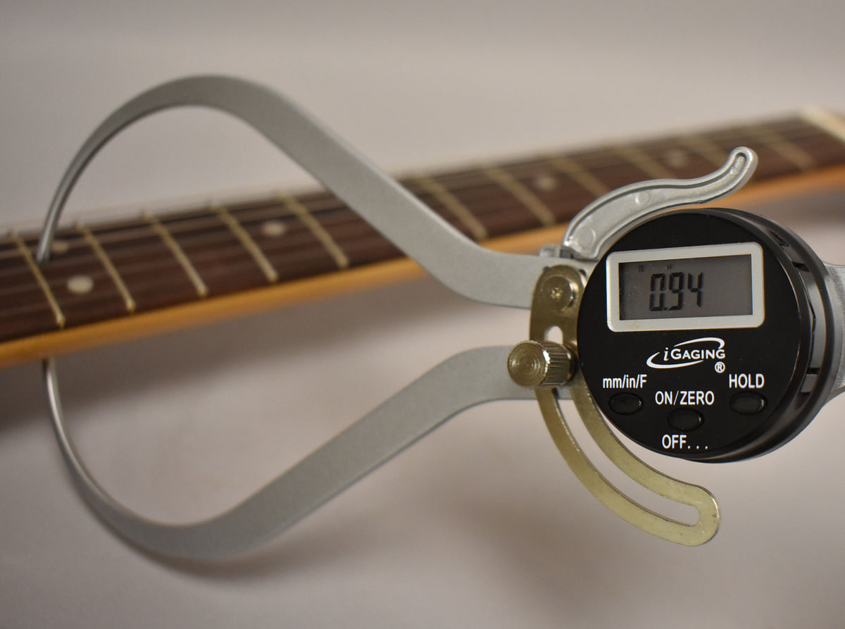 A digital caliper measures the string height at the 12th fret of the dark rosewood fretboard with white dot inlays.