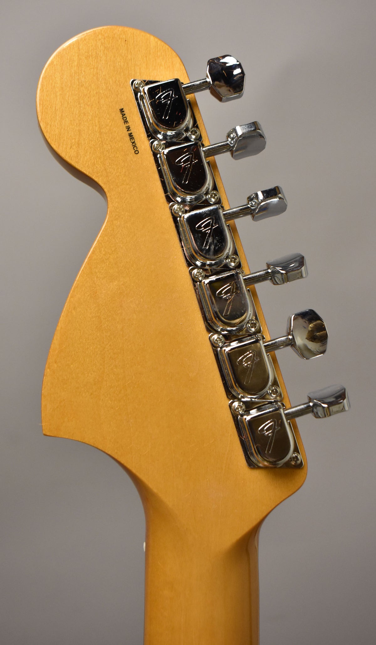 The back of a light brown wooden headstock and neck, angled right, with six chrome tuners and "MADE IN MEXICO" stamped vertically.