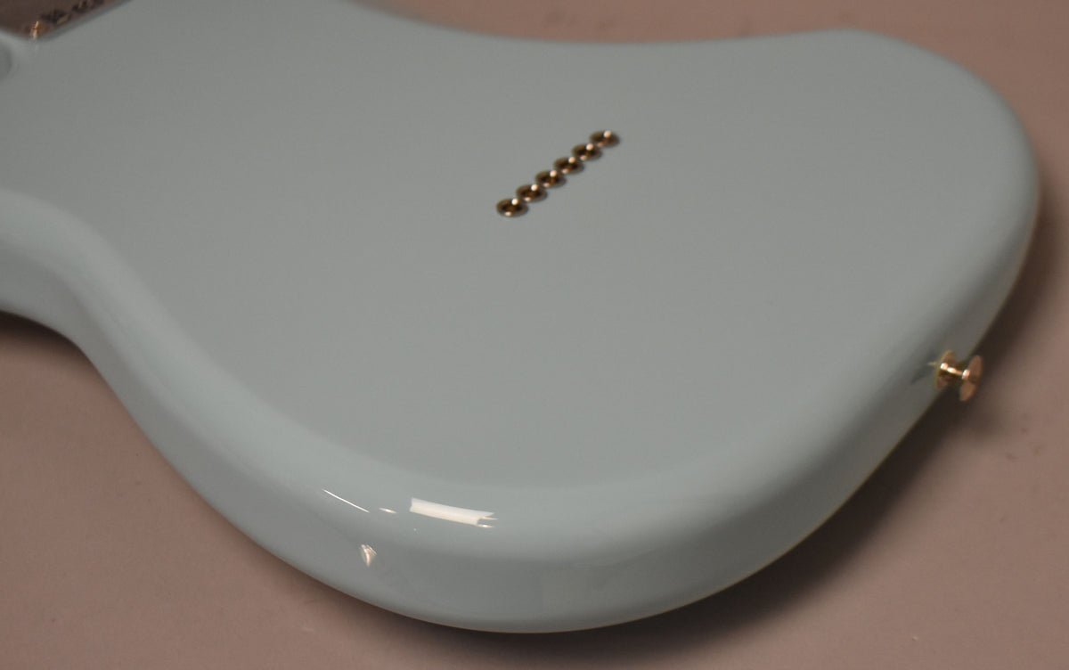 The back of a glossy Daphne Blue Stratocaster body, angled from the lower bout, showing six gold string ferrules and a gold strap button.