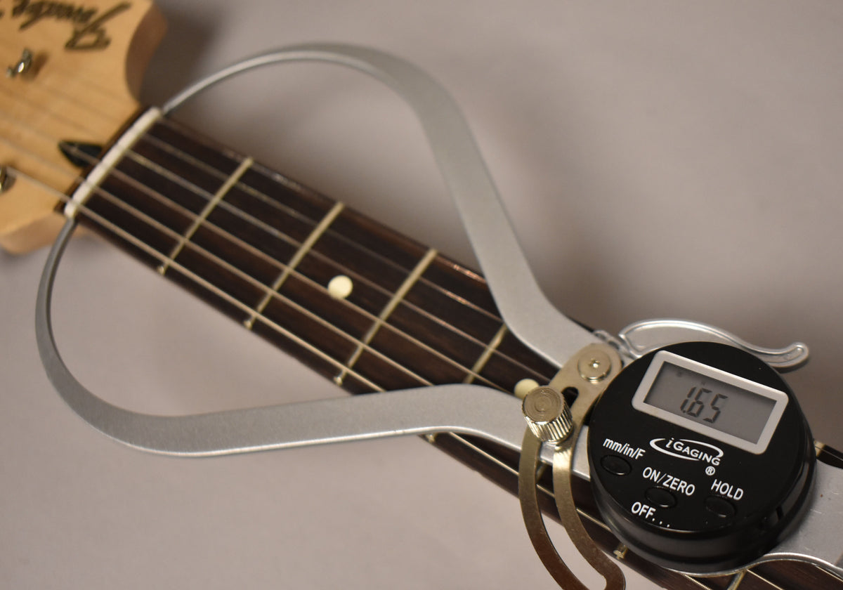 A close-up of the guitar's dark fretboard and light headstock, with a silver digital caliper measuring the nut width at 1.65 inches.