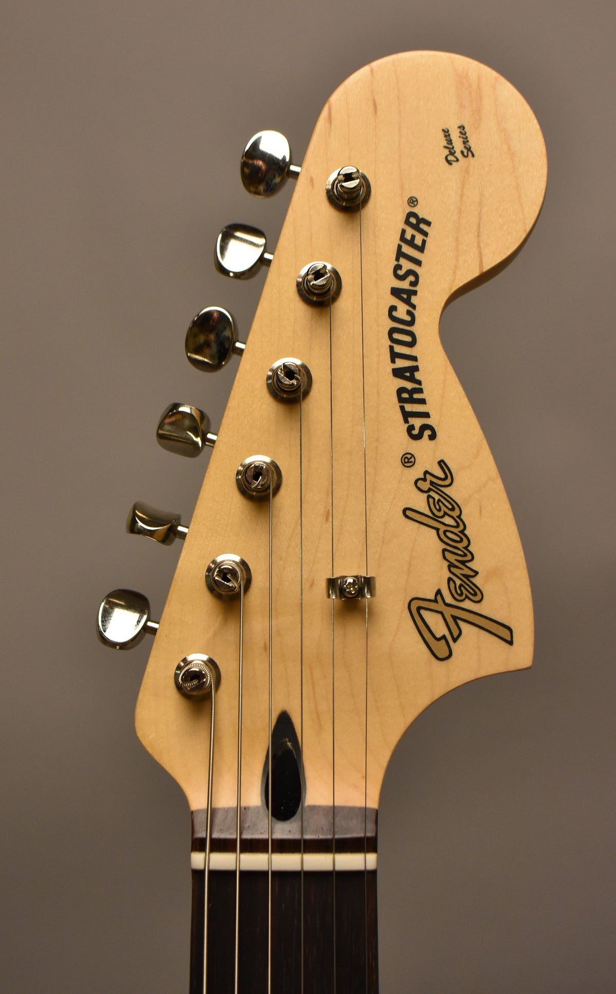 Close-up of a light wood guitar headstock with "Fender Stratocaster Deluxe Series" in black, six chrome tuners, and strings.