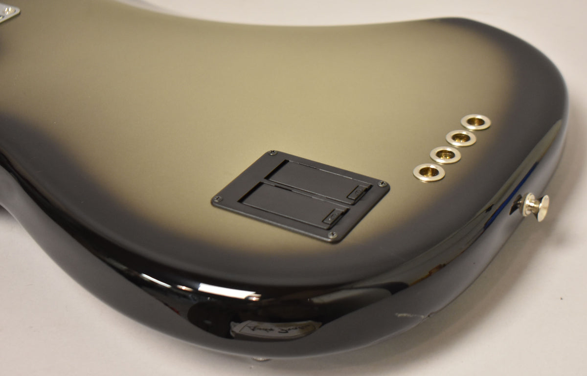 The bass guitar's silverburst body is angled, showing the back with a black battery compartment, four gold string ferrules, and a strap button.