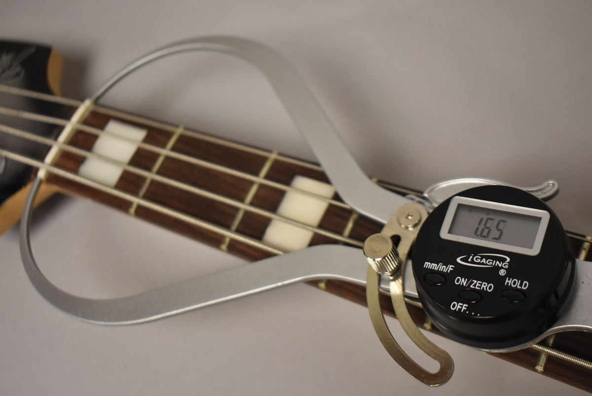 Close-up of the bass's dark fretboard with rectangular white inlays, showing a digital caliper measuring the nut width at 1.65.