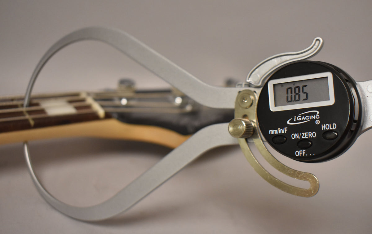 A digital caliper measures the bass's neck thickness at the first fret, displaying "0.85" on its screen.