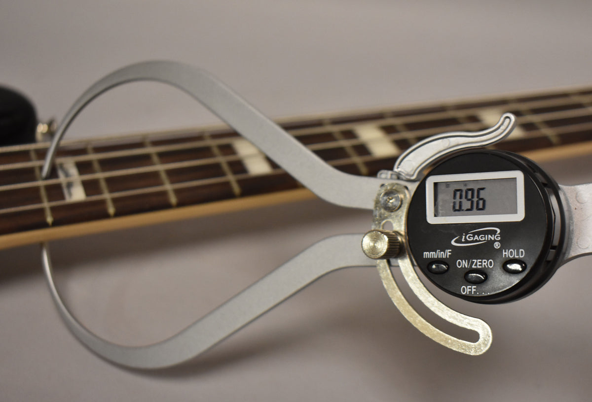A digital caliper measures the bass's dark fretboard and frets, displaying "0.96" on its screen.