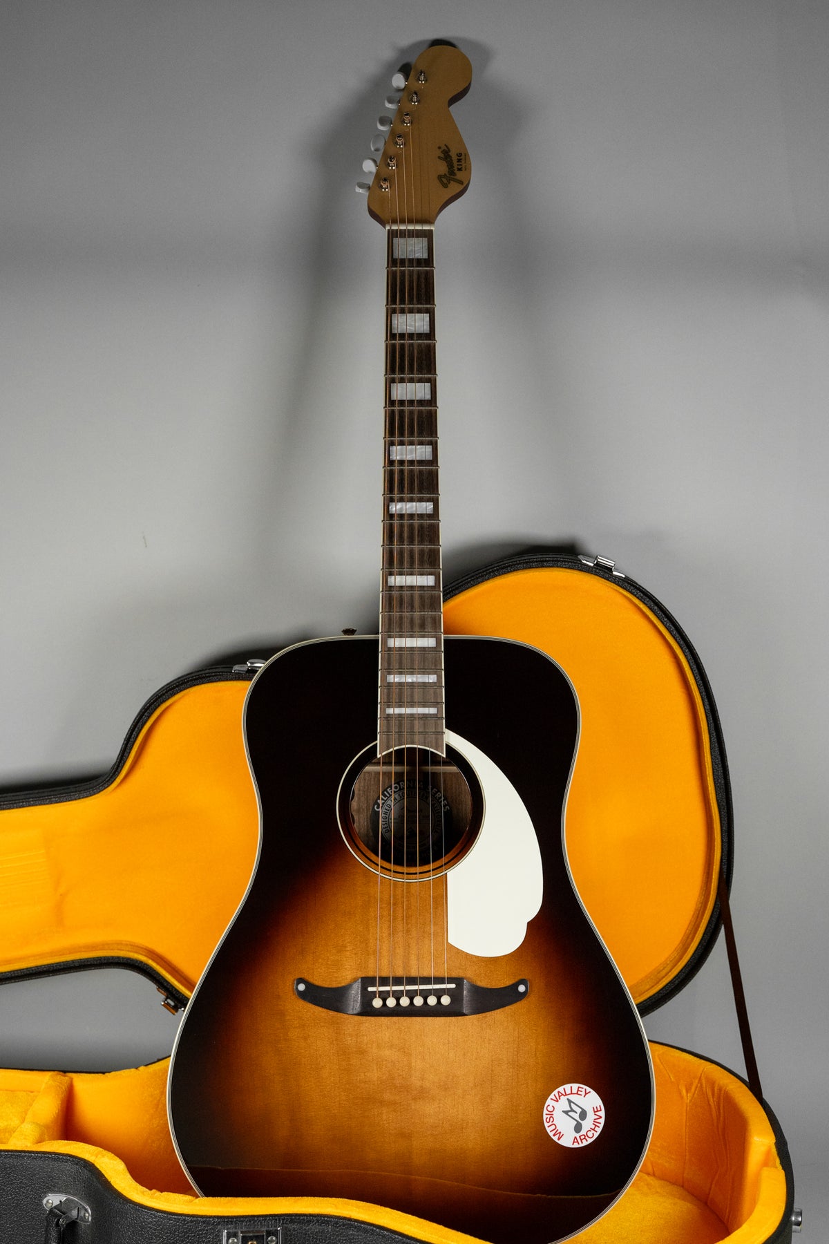 Full-length view of a sunburst acoustic guitar with a white pickguard, block inlays, and an open hard case with orange lining.