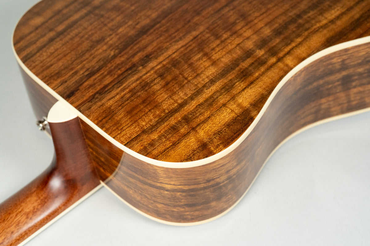 The guitar's glossy, dark brown back and side show prominent wood grain, with cream binding along the edges and a strap button visible.