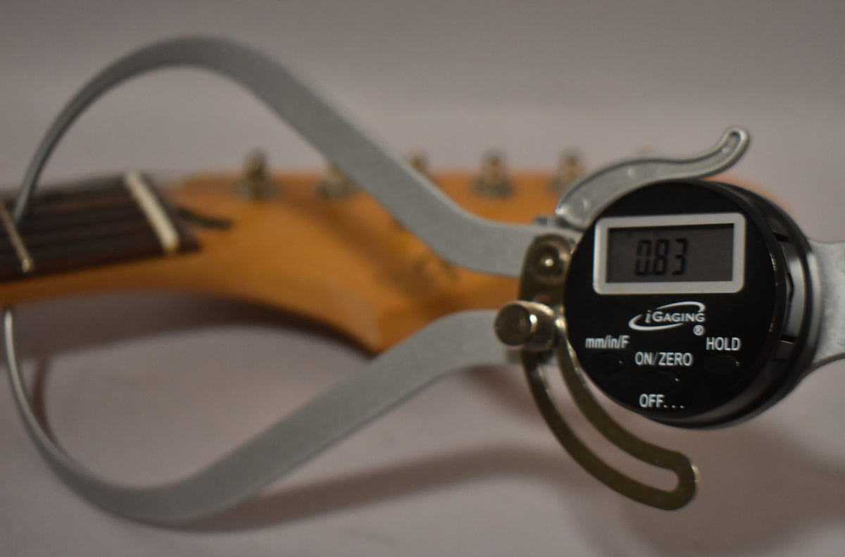 A digital caliper measures the guitar's neck thickness at the nut, displaying "0.83" on its screen, with the headstock blurred in the background.