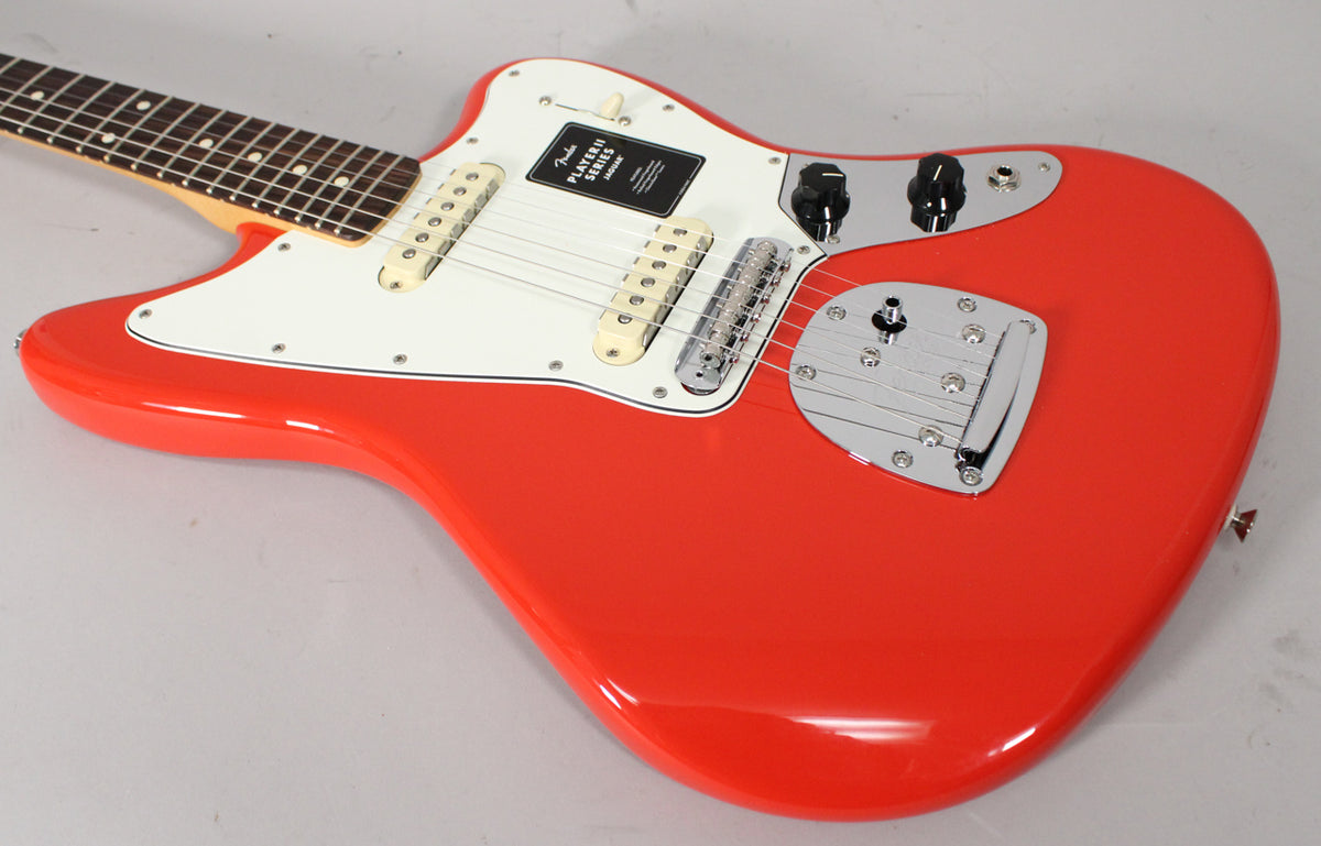 The coral red Jaguar guitar, angled from the neck, shows its white pickguard, two cream pickups, and chrome bridge and tremolo.