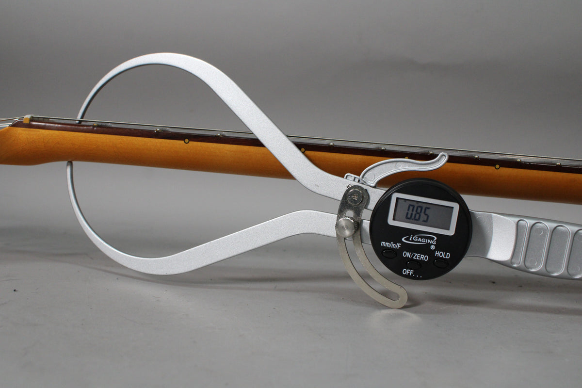 A digital caliper measures the neck thickness of the light brown guitar neck, showing 0.85 on its display.