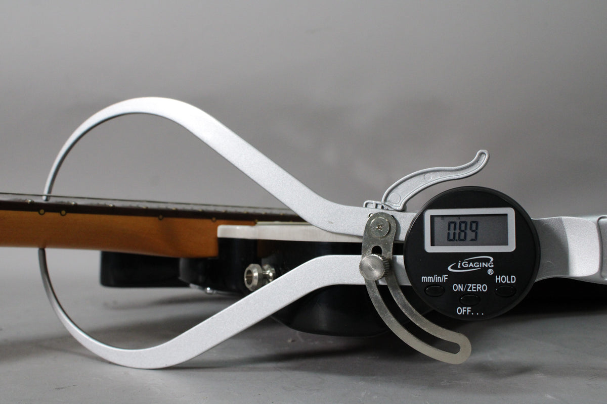 A digital caliper measures the guitar's neck relief, showing "0.89" on its screen, with the fretboard and body visible.