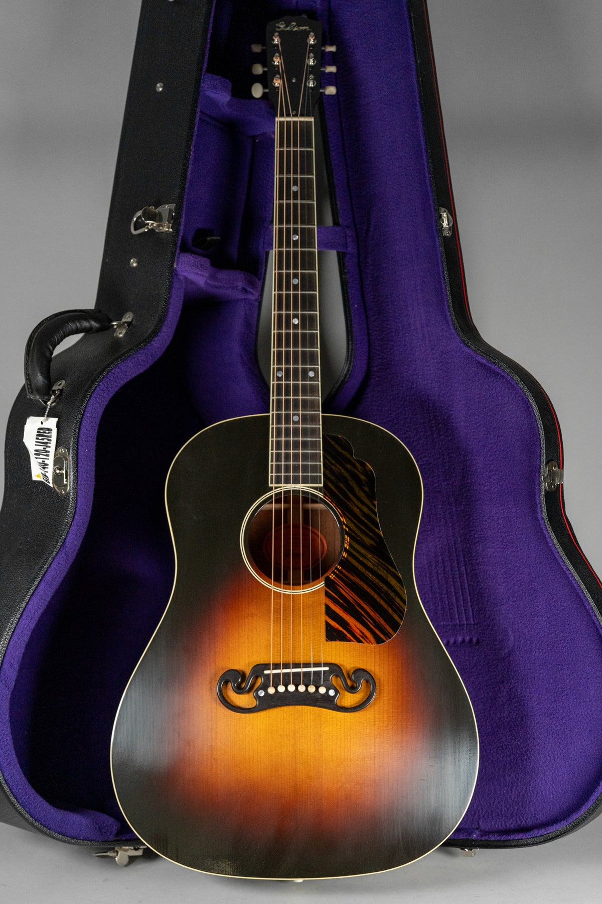 Full-length, front view of a sunburst acoustic guitar with a tiger-stripe pickguard, standing in its open, purple-lined hard case.