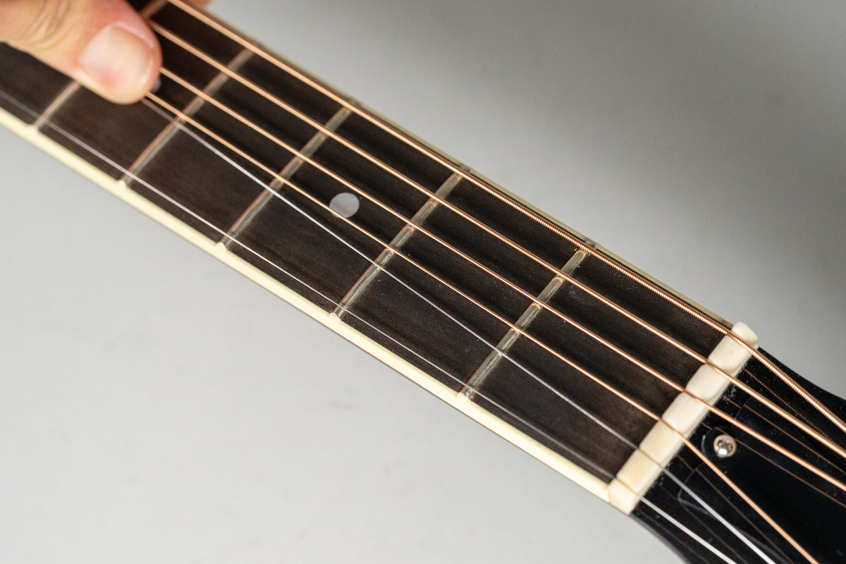 Close-up of the dark fretboard with light binding, showing the first five frets, a white nut, and a finger pressing a string.