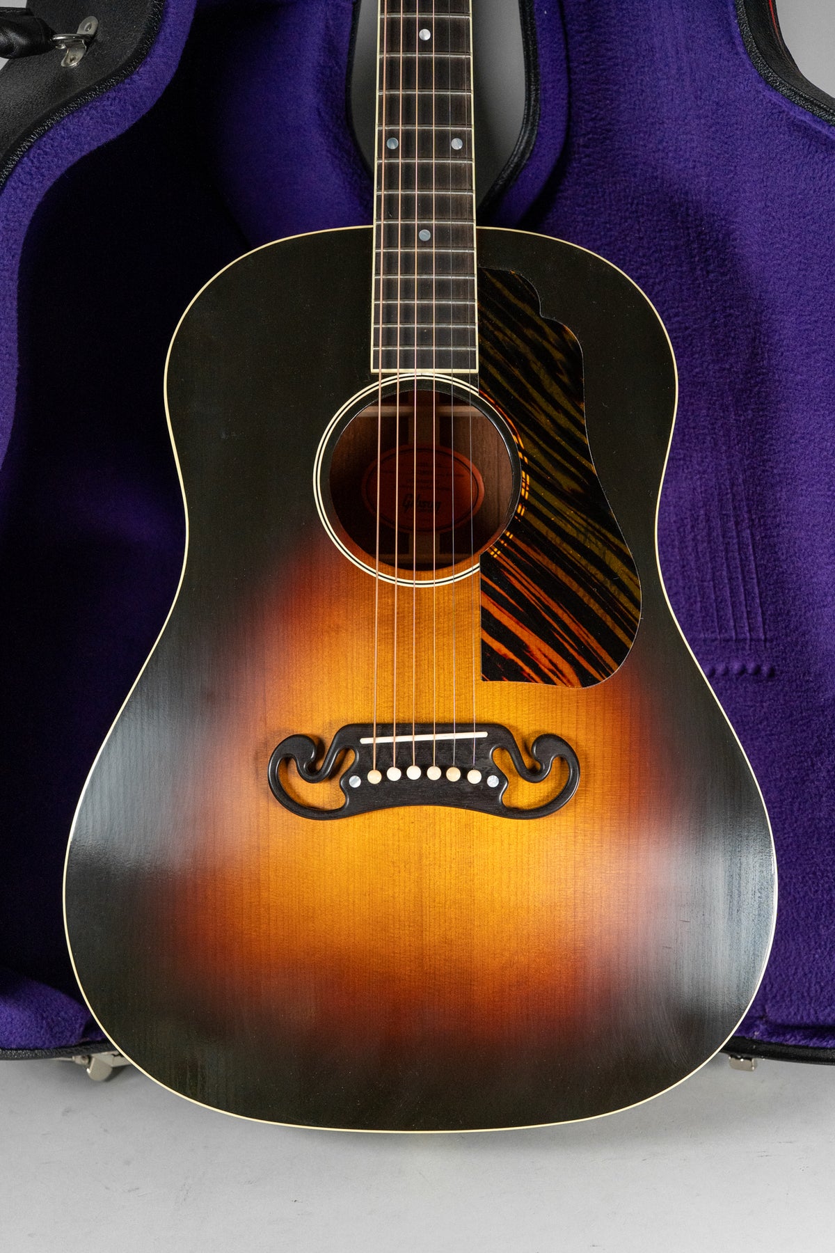 The acoustic guitar's sunburst body, with a tiger-stripe pickguard and mustache bridge, is framed against a purple case lining.