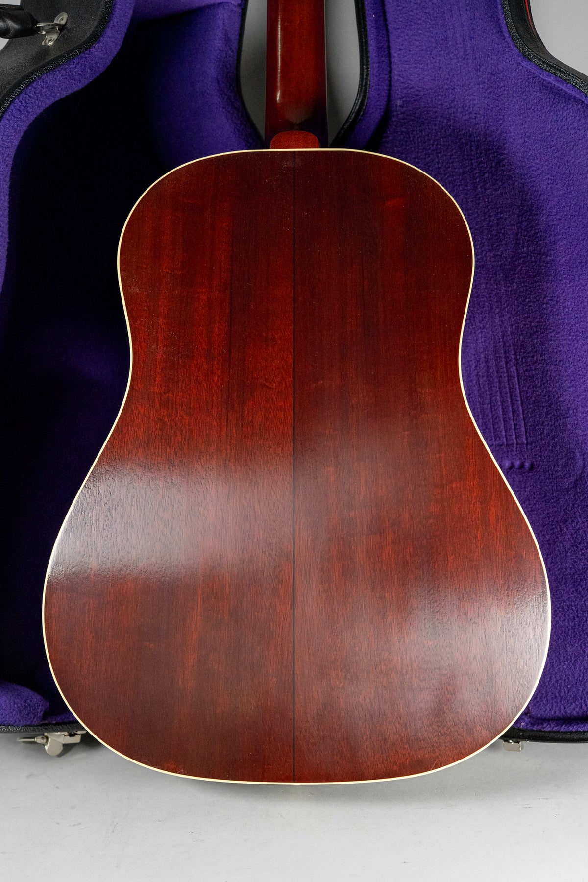 The guitar's dark reddish-brown back, with a light binding, is centered against a purple case lining and grey floor.