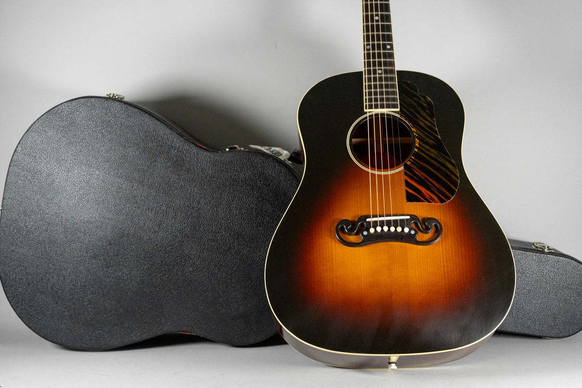 The acoustic guitar, with a dark sunburst finish and striped pickguard, stands next to its black textured hard case.