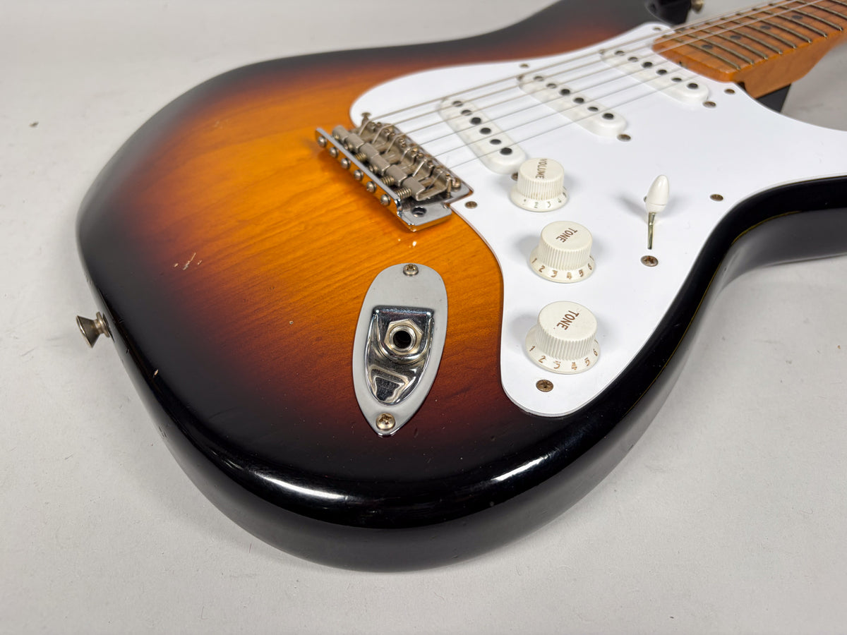 The lower body of a sunburst Stratocaster, showing the input jack, white pickguard, three cream knobs, and light wear.