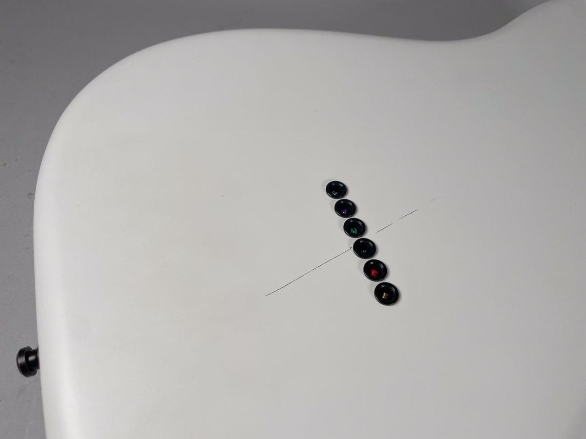 The back of a white guitar body, showing six black string ferrules in a diagonal line, with light scratches nearby.