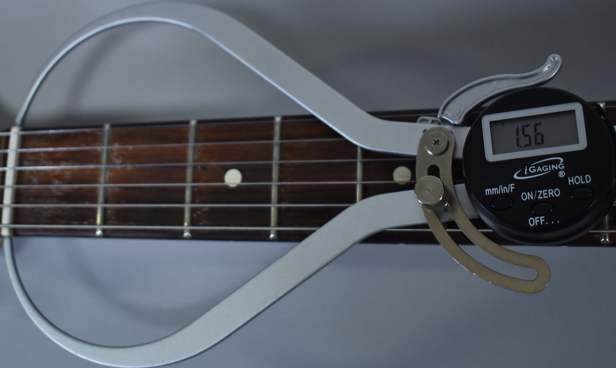 A digital caliper measures the guitar's fretboard, showing 1.56 on its display, with visible fret wear and dark wood grain.