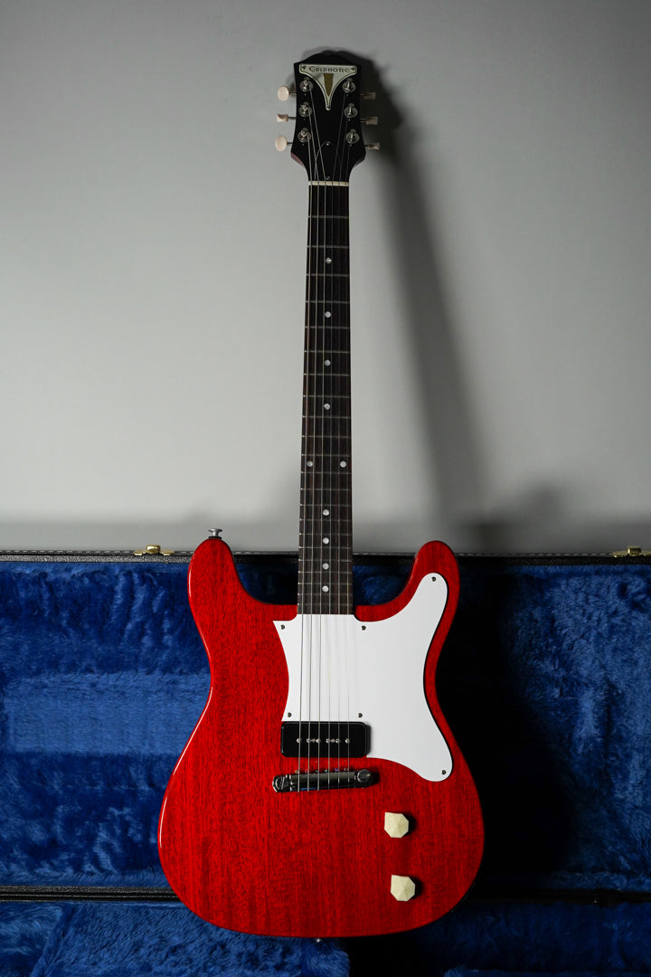 Full-length view of a cherry red solid body electric guitar with a white pickguard, a single black P90 pickup, and two cream control knobs, standing in a blue velvet-lined case.