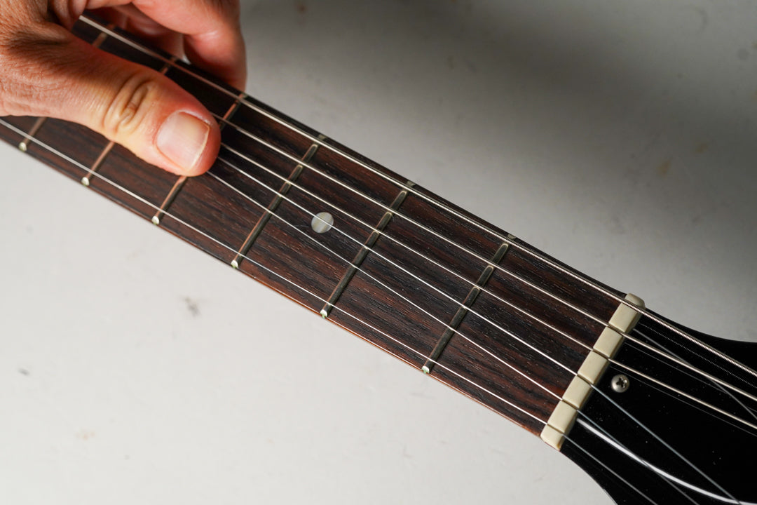 A hand frets the dark rosewood fingerboard with dot inlays, showing the cream nut and black headstock.