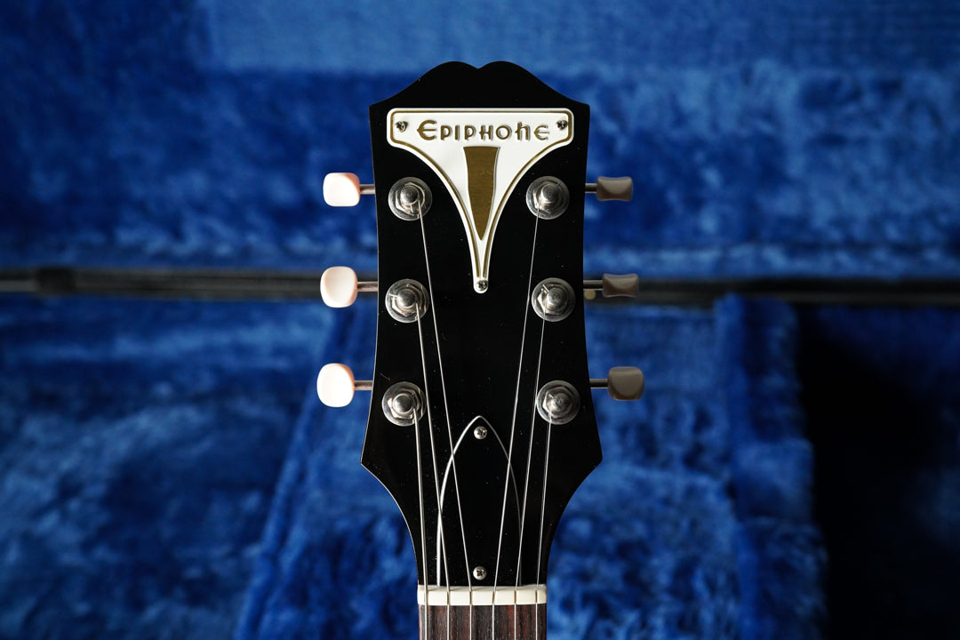 The guitar's black headstock with a white and gold inlay is centered, showing six tuners with cream buttons against a blue case lining.