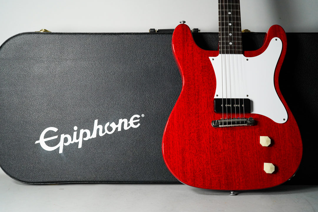 A cherry red solidbody electric guitar with a white pickguard and two cream control knobs, next to a black hard case.
