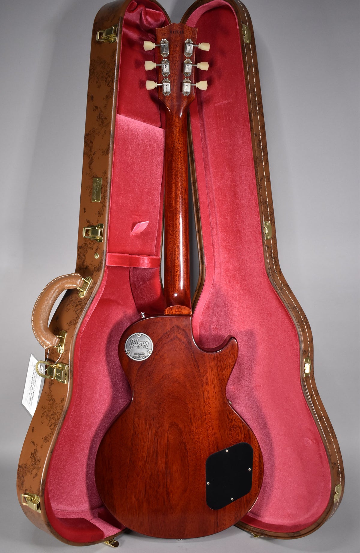 The guitar's dark cherry back and neck are visible, with a black control cavity cover, in a brown case with red plush lining.