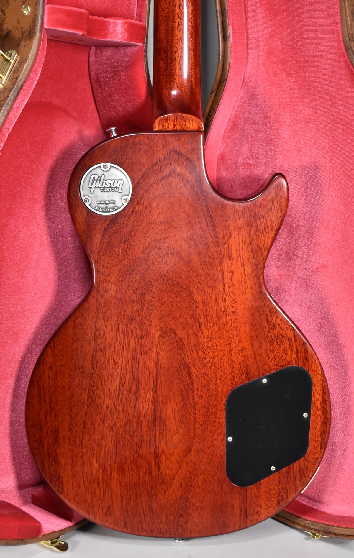 The guitar's mahogany back, with a visible wood grain, is shown in its open red-lined case, featuring a black control cavity cover.