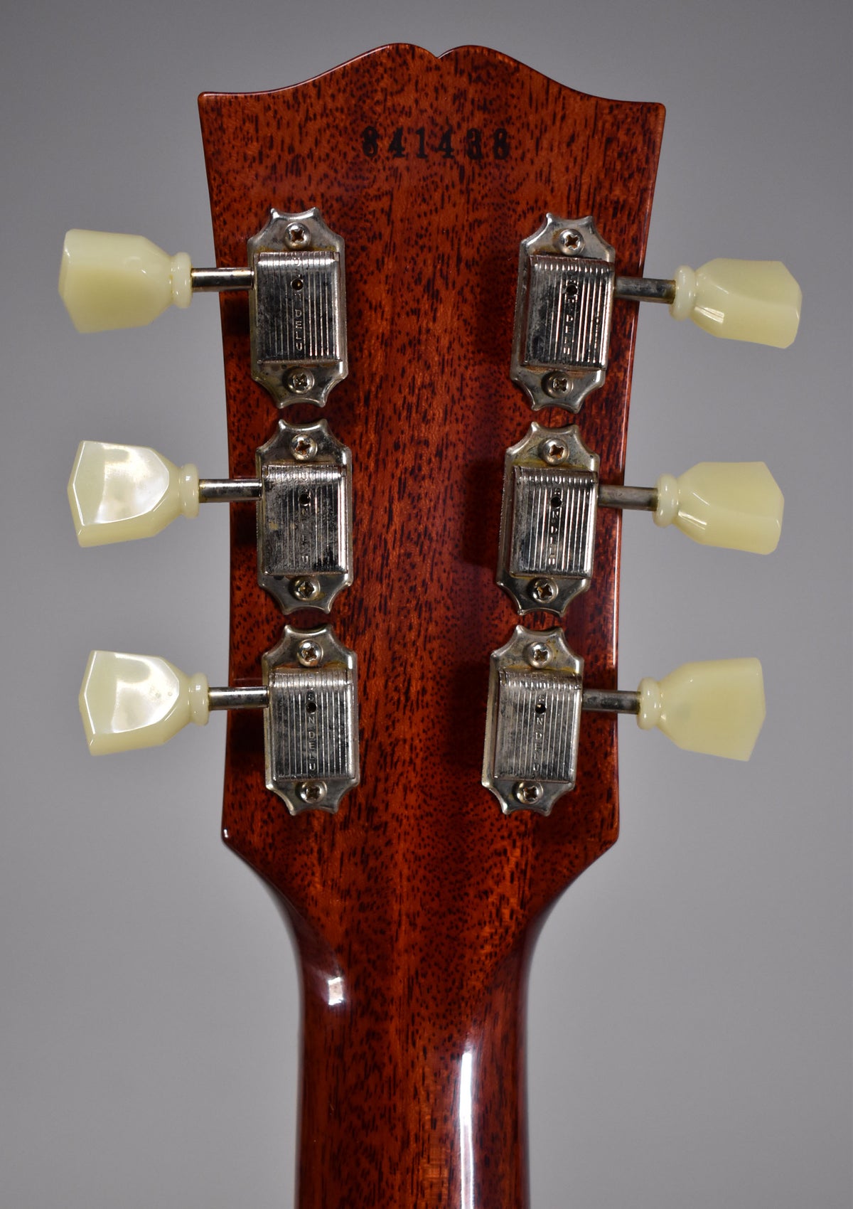 The back of a dark reddish-brown headstock with six cream tuning pegs and aged silver tuners, showing a serial number.