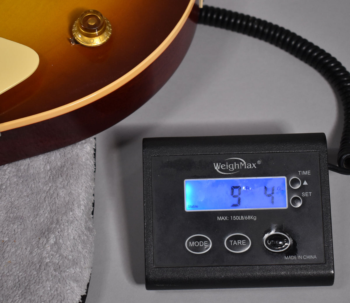 The guitar's iced tea burst body and cream binding rest on a grey towel, next to a scale displaying "9.4" lbs.