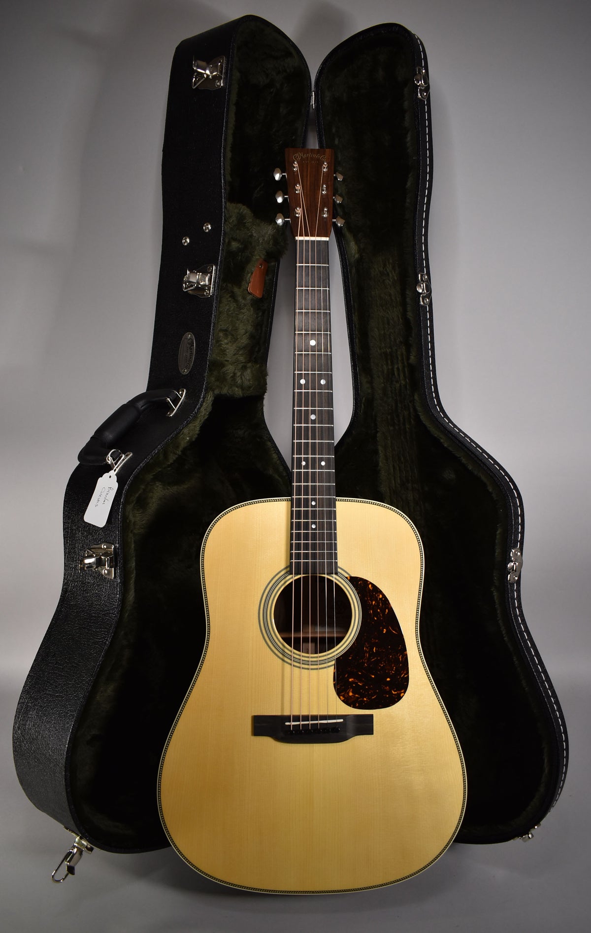 Full front view of a light-blonde acoustic guitar with a dark tortoiseshell pickguard, standing between its open, dark green-lined hard case.