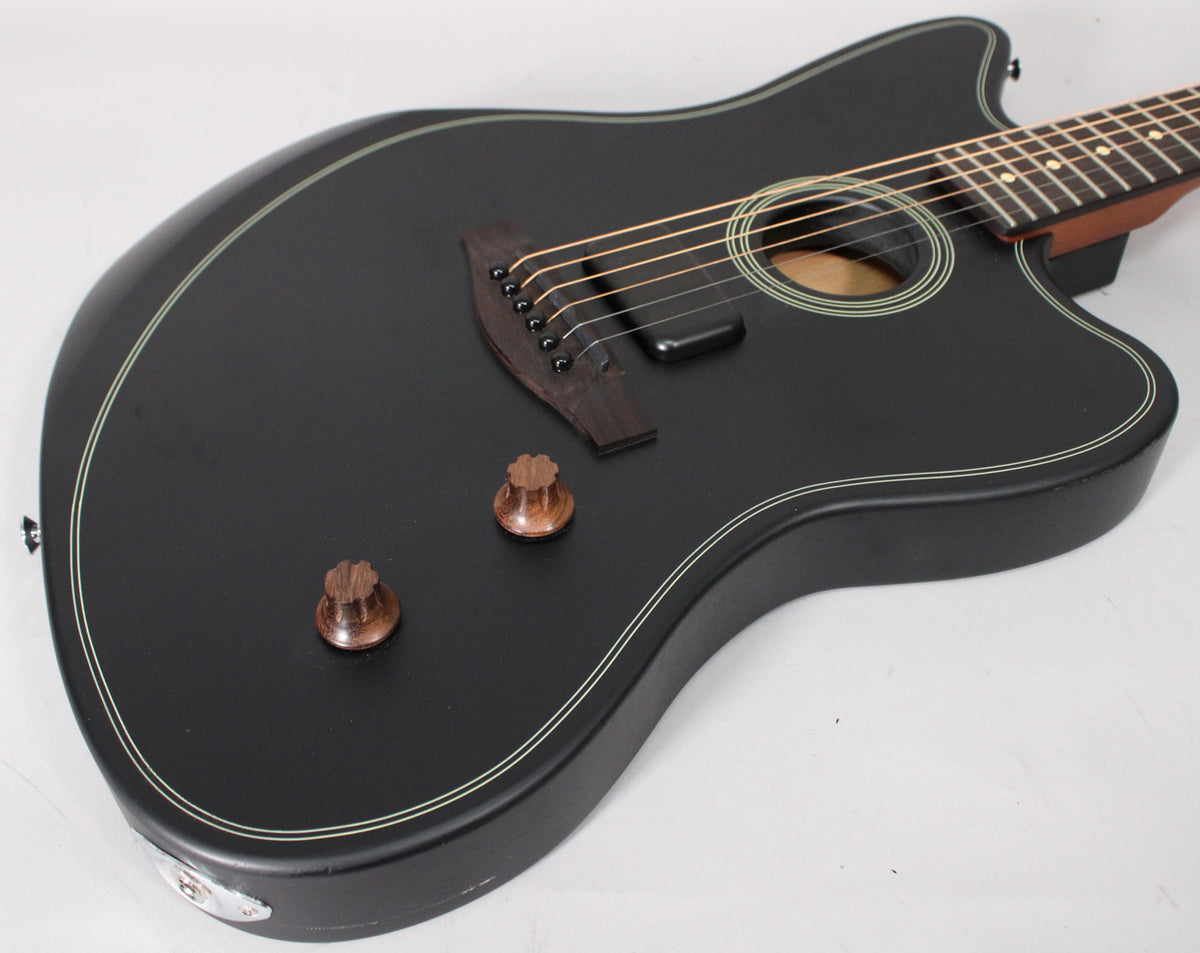 The matte black guitar body, with white binding, is angled from the lower left, showing two wooden control knobs and a soundhole.